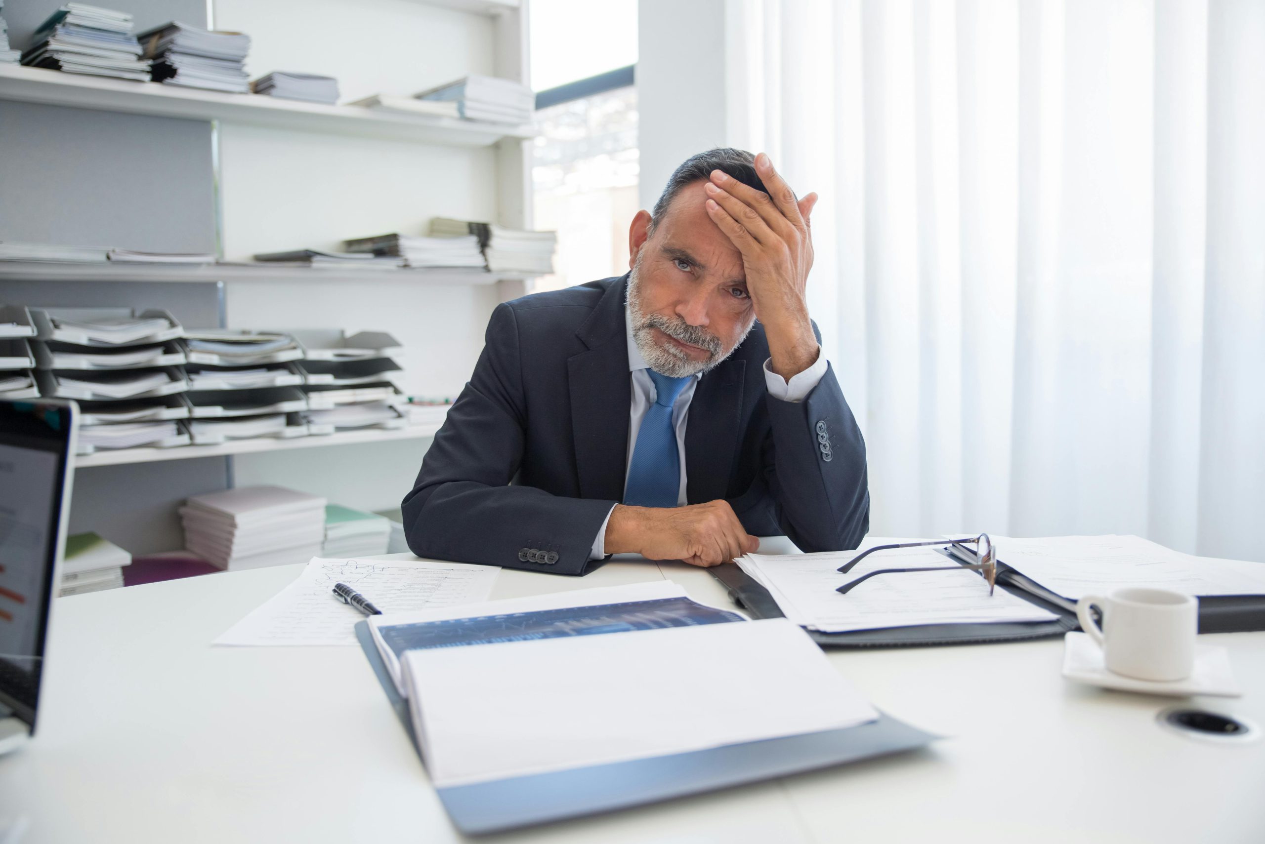 Elderly businessman in a black suit looking stressed at a desk with documents in a modern office.